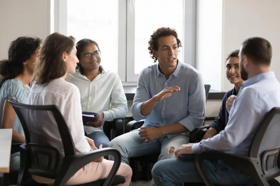 0x0 (19) A diverse group of colleagues sits in a circle in a bright office, engaged in a discussion with one person speaking.